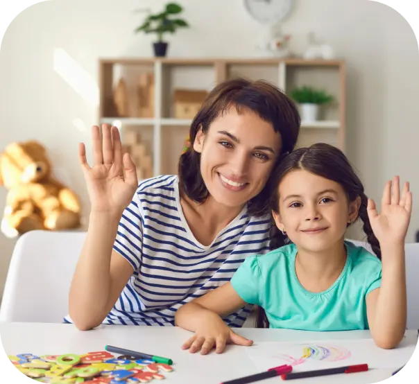 A woman and a young girl waving from a table with crayons, foam letters, and a drawing